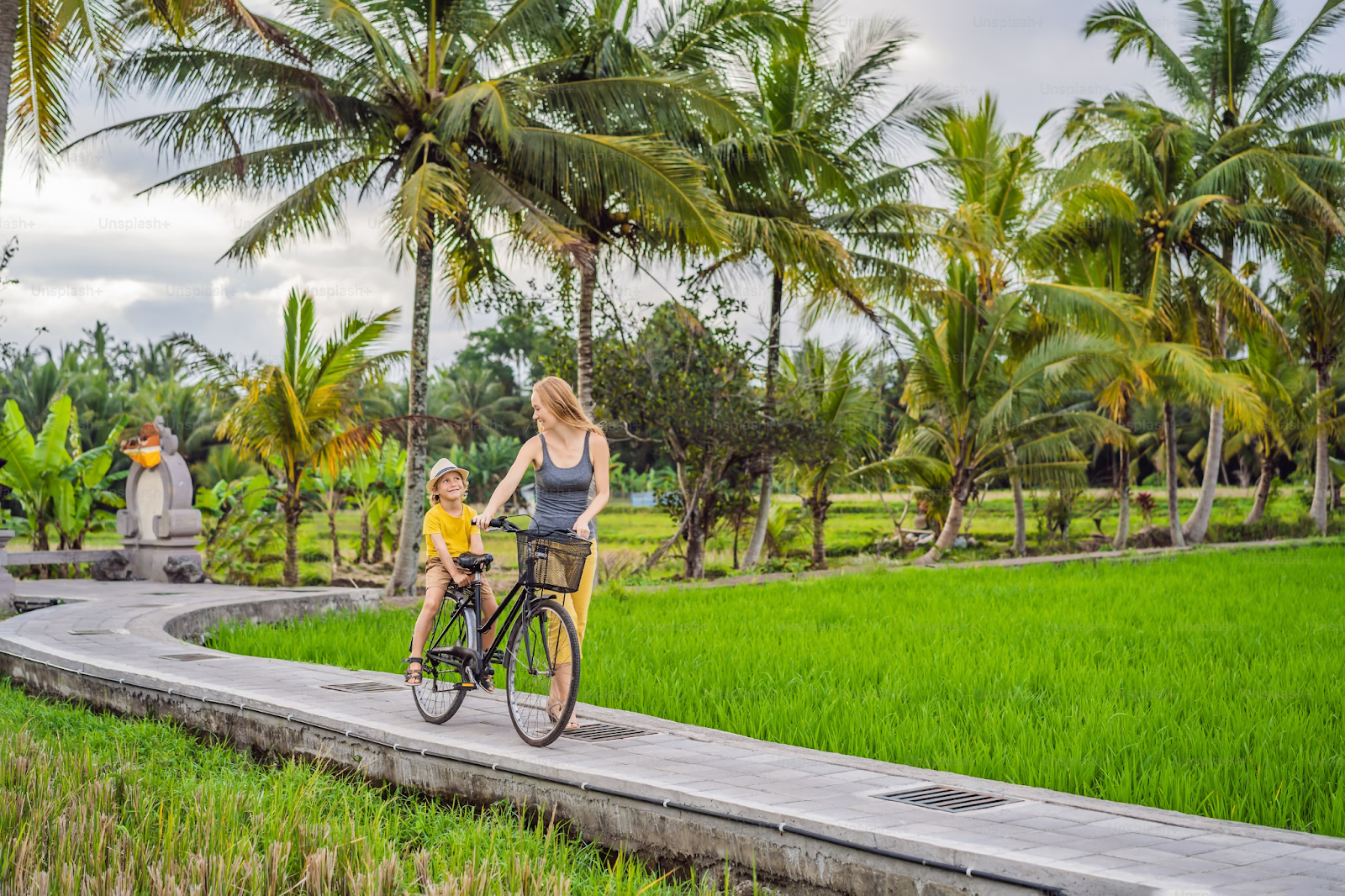A mother and her young child are riding a bicycle along a scenic path surrounded by lush green rice paddies and palm trees in Bali.