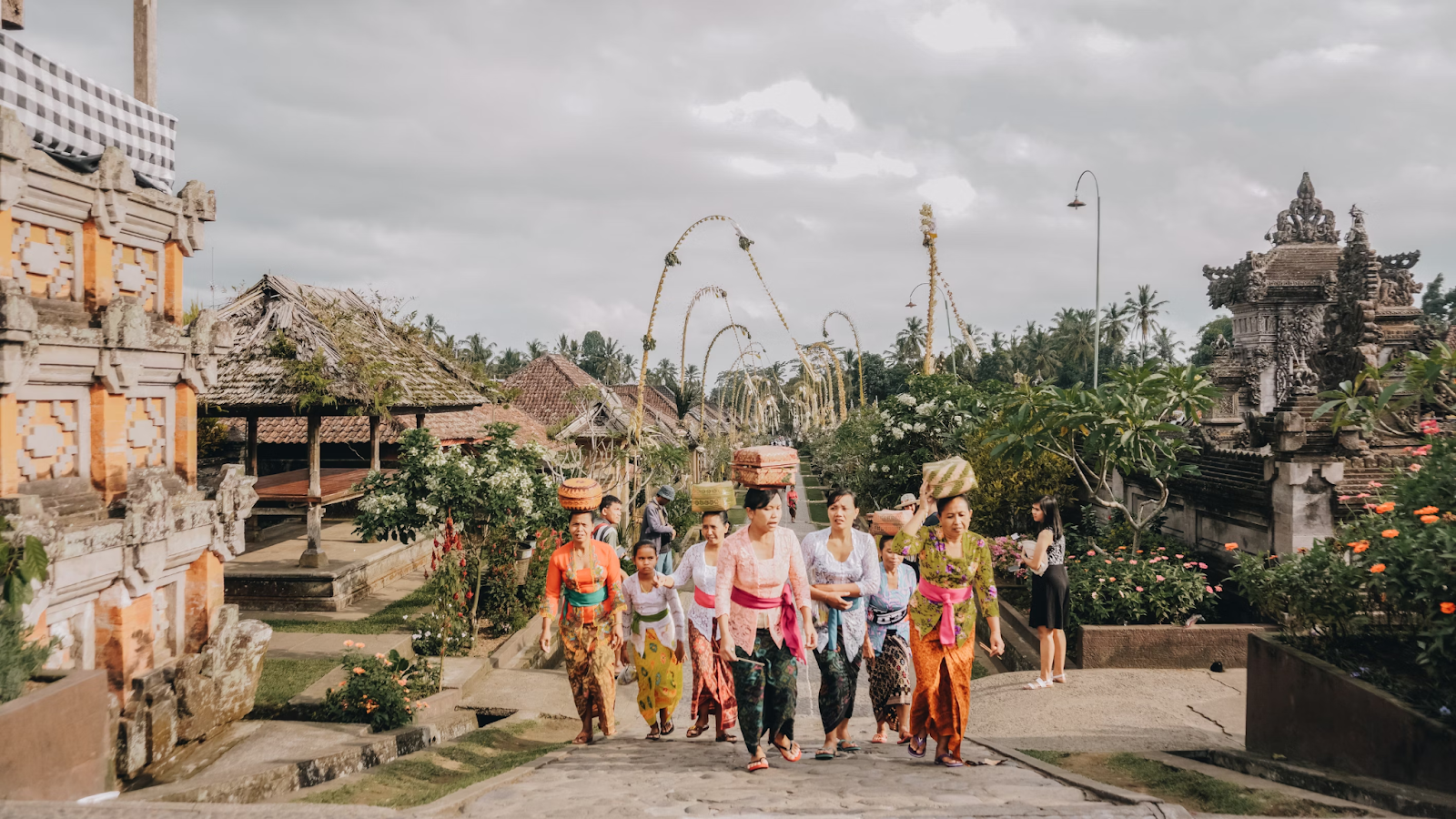 A group of Balinese women dressed in traditional attire, carrying baskets on their heads, walk through a village adorned with ceremonial decorations. The scene captures the cultural richness and vibrant community life of Bali, Indonesia, set against a backdrop of traditional Balinese architecture and lush greenery.