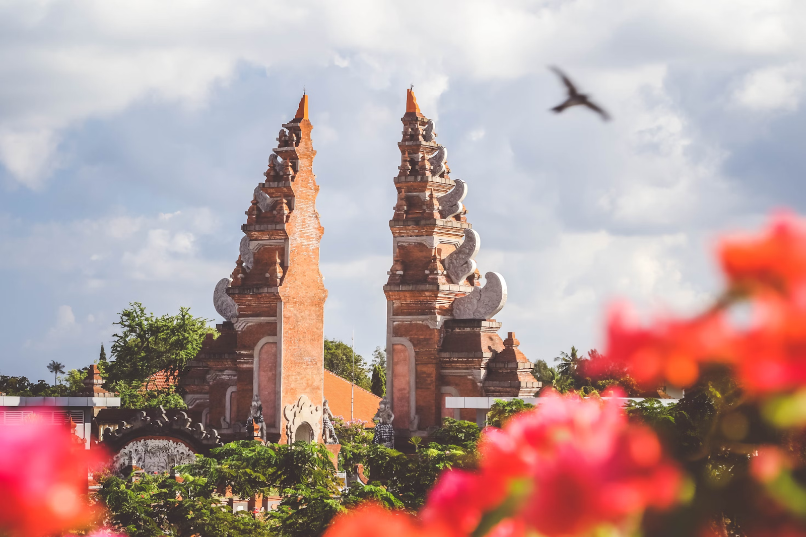 A traditional Balinese gate stands majestically against a backdrop of a cloudy sky. The twin red-brick structures are adorned with intricate carvings and floral decorations. In the foreground, vibrant pink flowers blur into the frame, adding a splash of color to the scene.